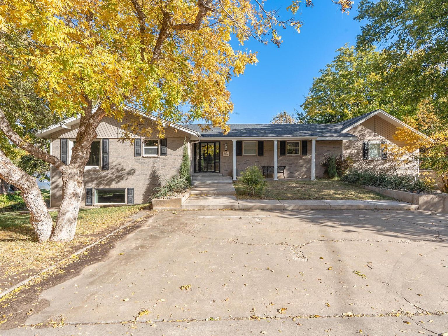 Front elevation of 147 S Shore Dr: a low single-story ranch with brick and pale-painted siding, mature trees turning autumn yellow on either side, broad driveway approach.