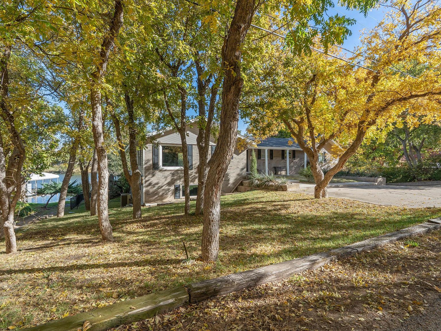 View of the residence at 147 S Shore Dr through a stand of yellow-leafed autumn trees on the shoreline approach, with a sliver of lake water visible at left.
